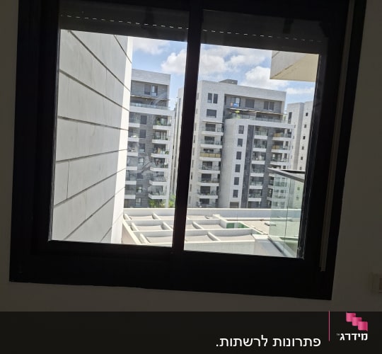 חלון אלומיניום כהה עם רשת נגד חרקים מבעדו נראים בניינים